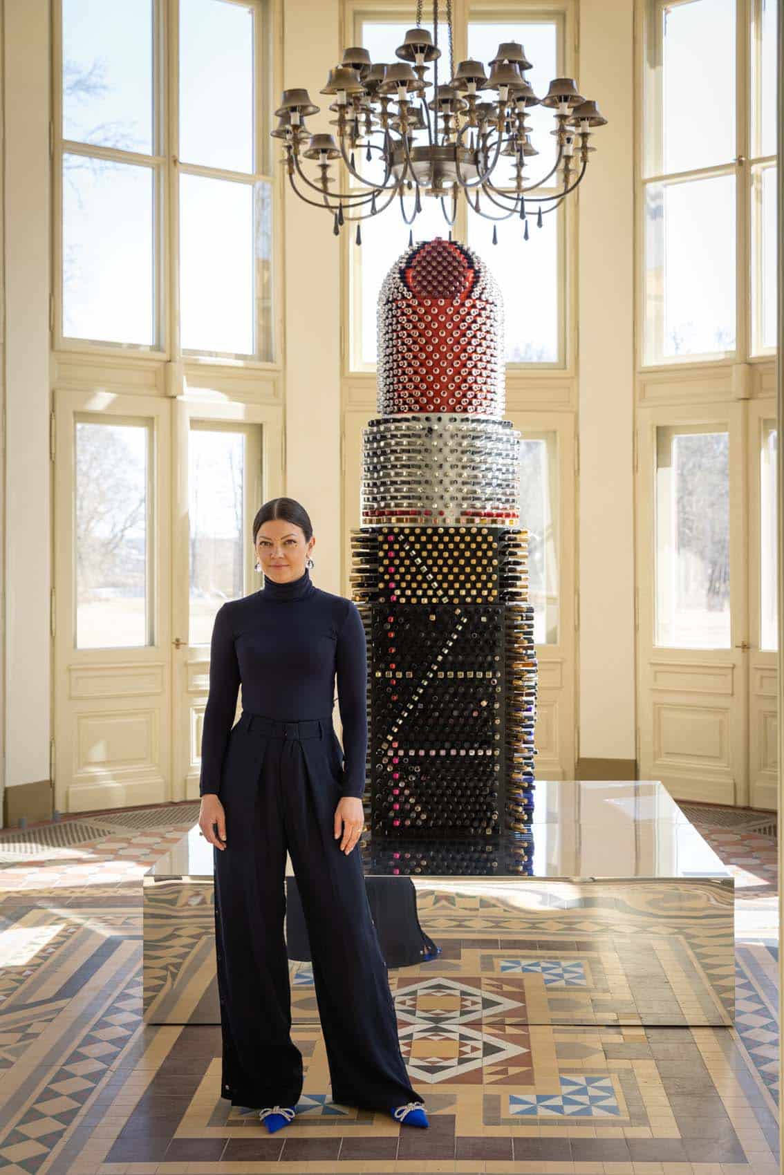 Giant Lipstick sculpture exhibited in interior with chandelier and large windows.