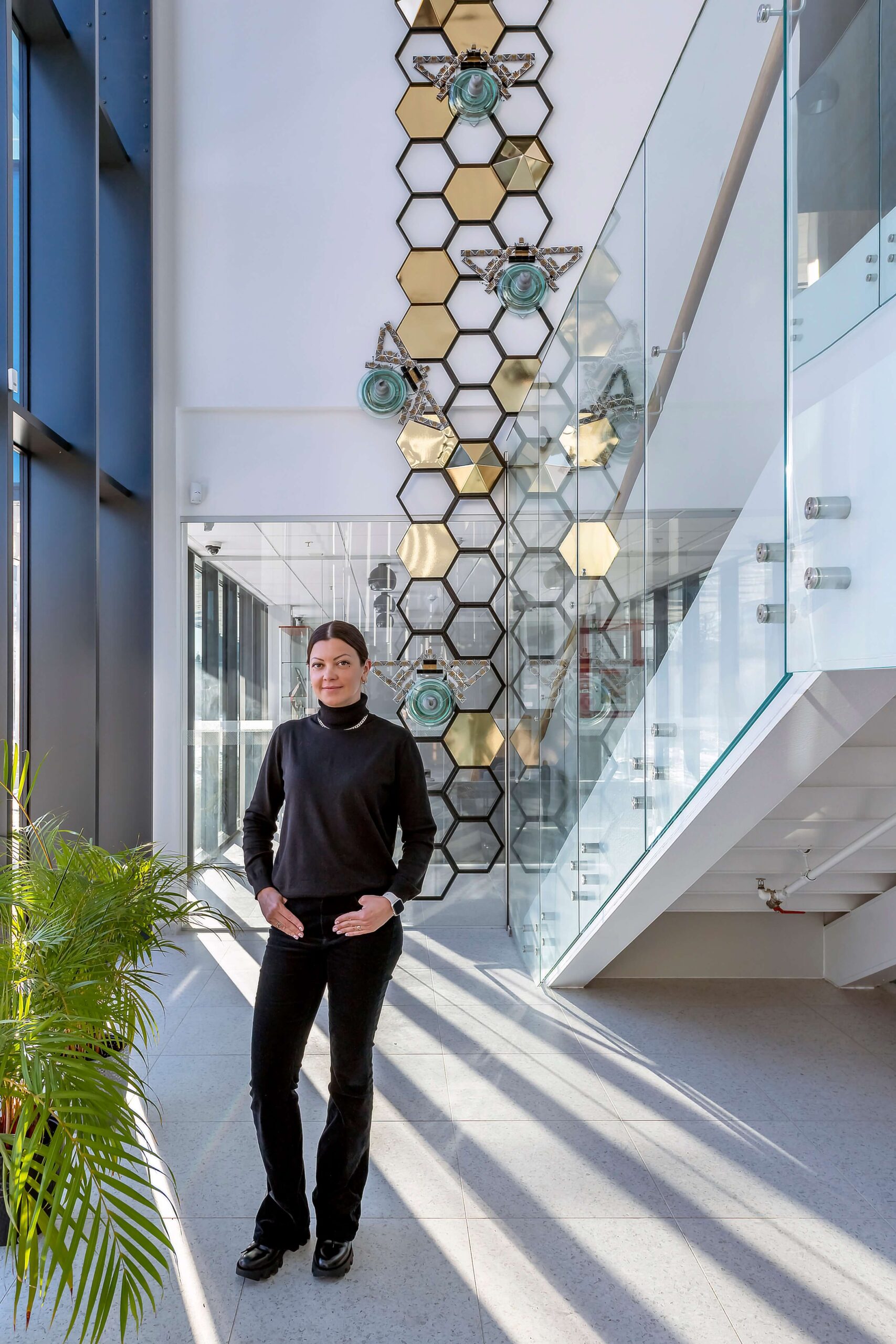 Artist Agne standing in front of her monumental "Honeycomb" upcycled art installation. The tall geometric piece features giant bees made from repurposed glass insulators climbing a gold and black honeycomb structure, displayed in a bright, modern building with a glass staircase.