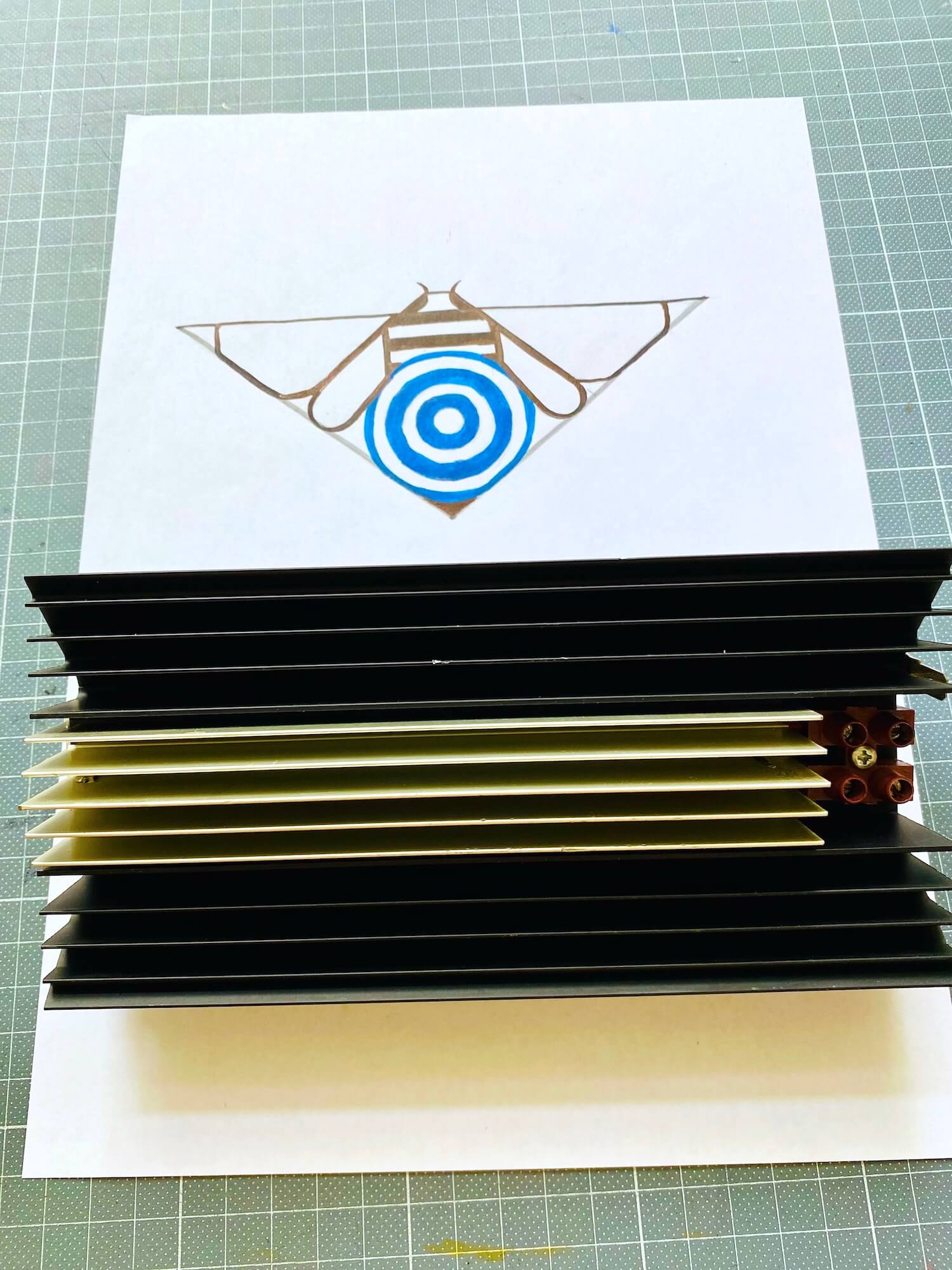 A black and gold electrical component placed over a colored drawing of a geometric bee, showing how the raw materials dictate the Art Deco aesthetic of the artwork.