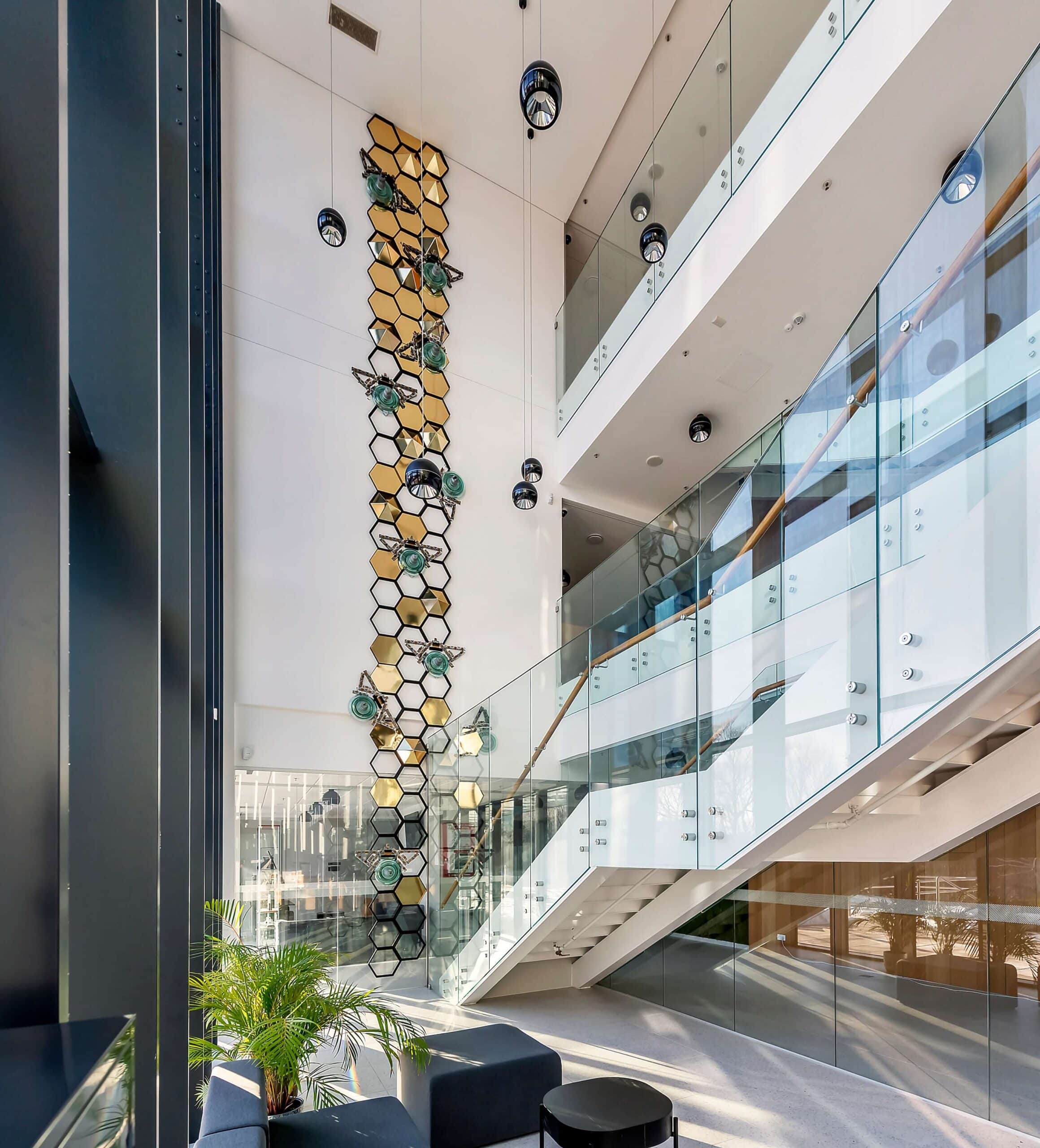 A ground-level view of the "Honeycomb" art installation ascending a tall white wall next to a modern glass staircase. The artwork features large bees crafted from industrial electrical waste climbing a vertical arrangement of gold and black hexagons, illuminated by natural sunlight.